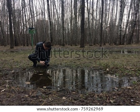 A young guy with brown hair in a leather jacket takes a picture of a large puddle in a park with bare trees on a cloudy autumn day.