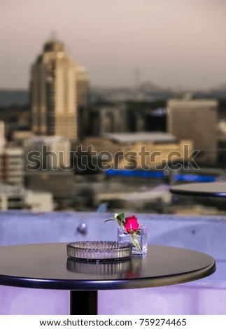 A flower in a small vase next to an ash tray on a table with a view of the city