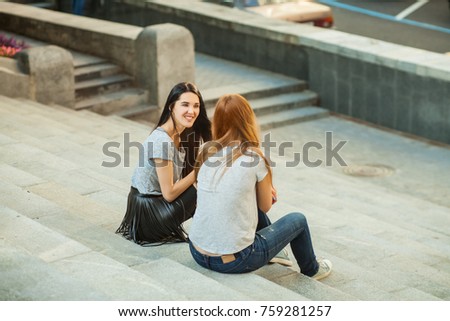 Two cheerful girls talking and sitting on the stairs in the street. one girl with long brunette hair in black skirt, another redhead girl in gray shirt and blue jeans. concept of sincere friendship Royalty-Free Stock Photo #759281257