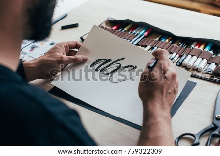 Calligraphy artist or student practices spelling words and letter with black ink brush on white canvas, sits behind table with different kinds of tools, pens, brushes and watercolors set up Royalty-Free Stock Photo #759322309