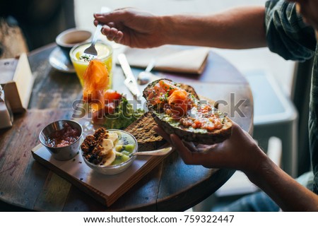 Young man enjoys big and tasty breakfast at downtown cafe, prepares delicious sandwich or toast with avocado spread and smoked salmon on top. chia seeds with yoghurt for dessert  Royalty-Free Stock Photo #759322447