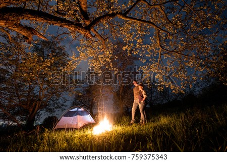 Romantic couple tourists standing at a campfire near tent, hugging each other under trees and night sky. Night camping