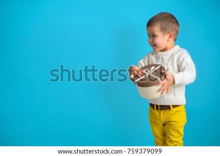 A happy smiling boy in a white sweater and yellow jeans is holding a  gift. Child waiting when you can open of a cardboard box tied with a ribbon. Isolate on  azure background. Christmas conceptual ph