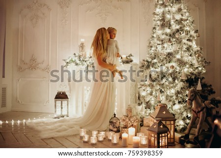 beautiful woman in a white dress in the floor holds a daughter in her arms is standing in a beautiful bright room with decoration near shiny Christmas tree and fireplace with garlands lanterns,candles