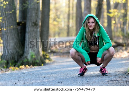 Female sport fitness model outdoors on forest path squats while wearing sweatshirt hoodie.