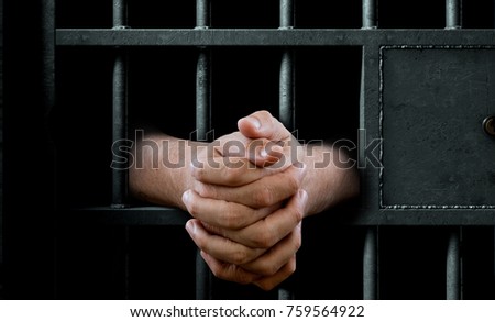 A closeup of a dimly lit prison holding cell door with arms reaching out in a clenched prayer position - 3D render Royalty-Free Stock Photo #759564922