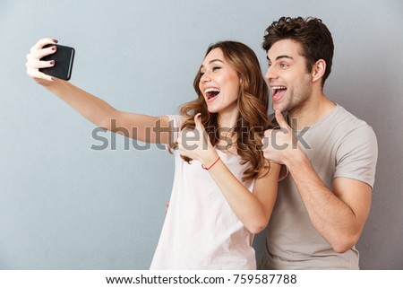 Portrait of a cheerful young couple showing thumbs up gesture while standing and taking a selfie over gray wall