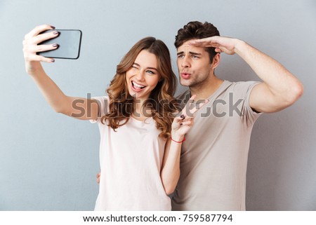 Portrait of a happy young couple showing peace gesture and winking while standing and taking a selfie over gray wall