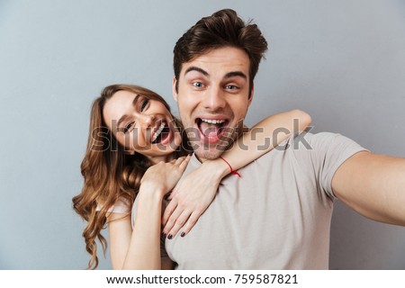 Portrait of an excited young couple hugging while standing and taking a selfie over gray wall
