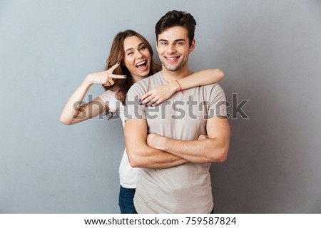 Portrait of a happy young couple hugging while standing and showing peace gesture over gray wall