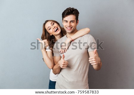 Portrait of a happy young couple hugging while standing and showing thumbs up gesture over gray wall