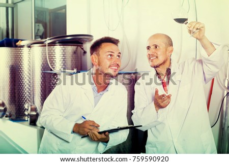 Two smiling workers wearing coats standing with glass of wine in fermenting section