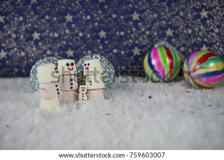 Christmas photography image using marshmallows shaped as snowman with icing for the smile and placed in snow with colorful bauble decorations and star pattern background