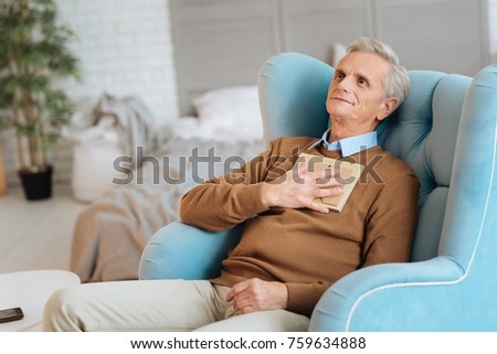 I will keep our love alive. Happy retired gentleman relaxing in a chair with a framed photo on his chest and looking into vacancy while thinking about his wife.