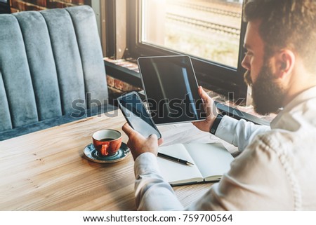 Back view.Young bearded businessman in white shirt is sitting at table,using smartphone,tablet computer,working,studying.On table is tablet computer,cup of coffee.Online marketing,education,e-learning