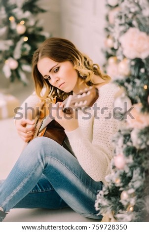 young woman in white sweater holds a guitar in hand and sits in christmas studio 