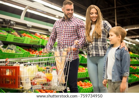 Portrait of happy young family with child shopping for groceries in supermarket together, smiling while pushing shopping cart in aisle past vegetable row