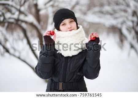 beautiful teenage girl in a black jacket and hat, with a white scarf, is holding two red Christmas-tree toys in her hands.  A child in the background of a snow-covered city.