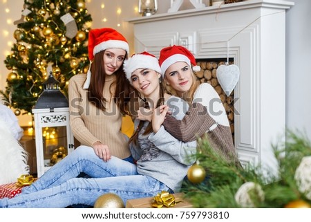 Picture showing group of beautiful girls in red christmas hats having fun near the fur tree in frot of the firplace, celebrating new year and hugging