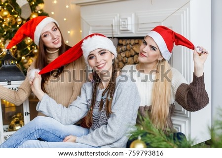 Picture showing group of beautiful girls in red christmas hats having fun near the fur tree in frot of the firplace, celebrating new year and hugging