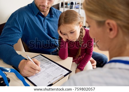 Portrait of frustrated little girl looking away with sad expression while visiting doctors office with father signing forms