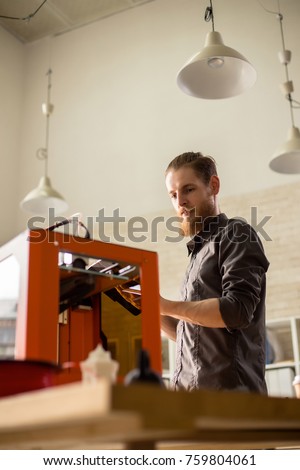 Low angle view of handsome bearded designer using 3D printer while working on ambitious project, interior of modern studio on background