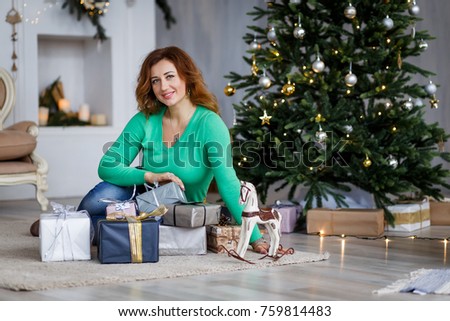 A beautiful woman with christmas present in front of the fur-tree and a fireplace with candles. New year's eve. Christmas eve. Cozy holiday at the fir-tree with lights and gold decor.