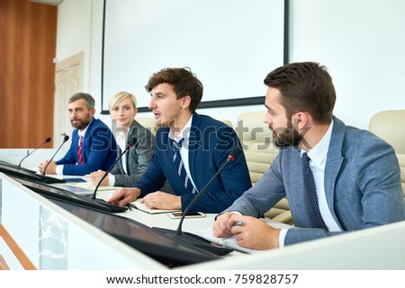 Portrait of several business people sitting in row participating in political debate during press conference answering media questions speaking to microphone Royalty-Free Stock Photo #759828757