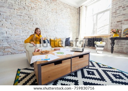 young beautiful woman is sitting on a sofa on a white brick wall background with a cup of coffee. Laptop, documents, drawings. Girl freelancer is resting.
