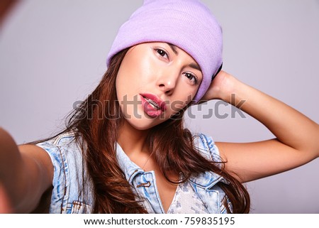 Portrait of beautiful cute brunette woman model in casual summer jeans clothes with no makeup in purple beanie making selfie photo on smartphone isolated on gray