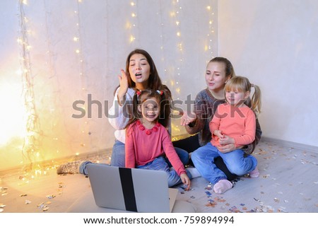 Two young and beautiful older girls together with two younger children of girls watch cartoons or movies on computer, communicate and laugh, sit on floor in bright room on background of wall with