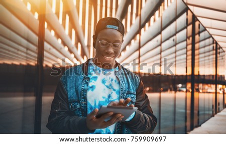 Handsome young black male student with white smile in glasses is checking his incoming messages via tablet pc; young handsome African guy waiting is chatting using digital pad near modern glass facade