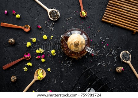 Brew the aromatic tea. Tea pot near wooden spoons with dried tea leaves, flowers and spices on black wooden background top view