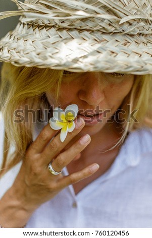 Smiling blond woman in a white shirt and traditional Balinese straw hat holding a Frangipani flower. Portrait picture. Tropical holidays. Happiness and healthy lifestyle. Bali, Indonesia.