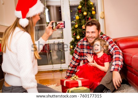 christmas, holidays, family and people concept - mother photographing happy father and little daughter with present by smartphone 