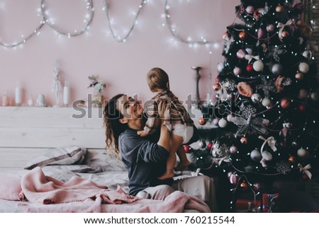 A young curly-haired mother in a gray sweater with a one-year-old daughter sits on the background of a Christmas tree by the pink wall in the Christmas and New Year's interior.
