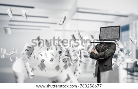 Cropped image of businessman in suit with laptop instead of head keeping arms crossed while standing among flying papers inside office building.