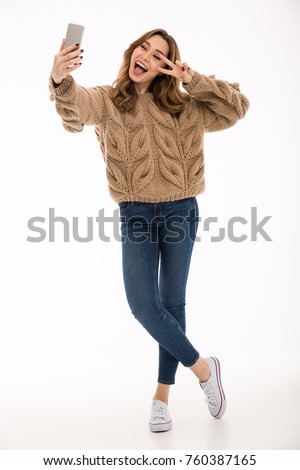 Excited cheerful young cute lady dressed in warm sweater standing isolated over white wall background. Looking aside make selfie by phone showing peace gesture.