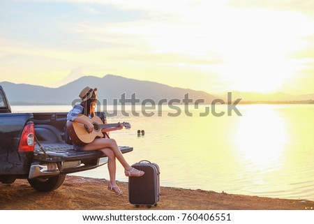 Asia traveler sitting hatchback car and playing guitar when sunset near the lake on holiday.beautiful tourist watch view of nature on vacation.