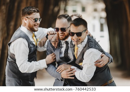 Groomsmen in black sunglasses hug groom tightly posing in the park Royalty-Free Stock Photo #760442533