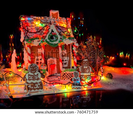 Adorable gingerbread family near big snow-covered homemade gingerbread house with Christmas lights on dark background. Mockup for seasonal offers and holiday post card
