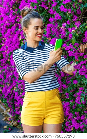 Colorful Freshness. smiling stylish woman in yellow shorts and stripy shirt against colorful magenta flowers bed with cellphone taking photo