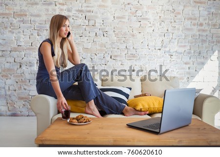 Female freelancer in her casual home clothing working remotly from her dining table in the morning. Houses on a sofa on a white brick wall background. Architect, blueprints. talking on the phone
