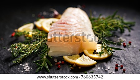 Fresh raw cod fillet with addition of herbs and lemon slices on black stone background
