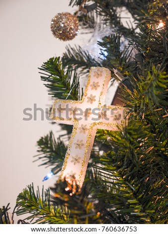 Closeup photograph of a beautiful homemade white and gold cross Christmas ornament hung from an artificial douglas fir Christmas tree lit with lights for a gorgeous religious holiday background