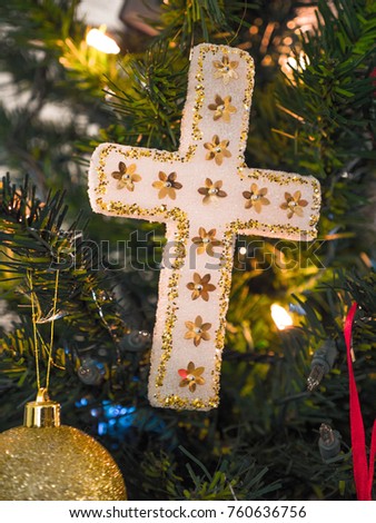 Closeup photograph of a beautiful homemade white and gold cross Christmas ornament hung from an artificial douglas fir Christmas tree lit with lights for a gorgeous religious holiday background