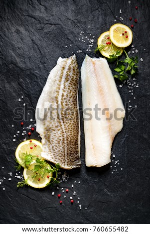 Fresh fish,  raw cod fillets with addition of herbs and lemon slices on black stone background, top view
