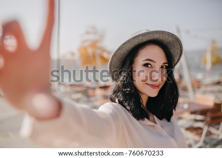 Dreamy brunette girl with short hair expressing happy emotions while making selfie outdoor. Photo of elegant young woman in hat and blouse taking picture of herself on blur background.