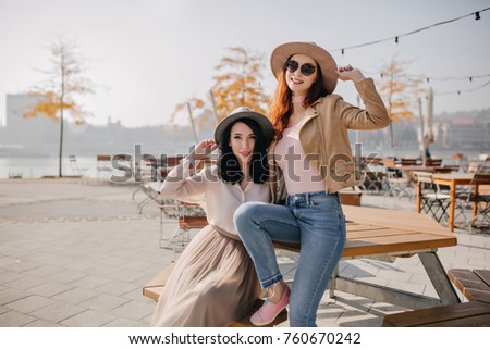 Ecstatic ginger woman in pink shoes sitting on wooden table in sunny day and laughing. Portrait of two adorable girls having fun in outdoor cafe not far from embankment.