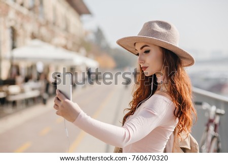 Charming red-haired woman with smartphone making selfie during walk in city. Serious ginger girl with trendy makeup taking picture of herself in hat on blur urban background.